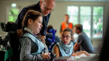 Ein Mädchen im Rollstuhl arbeitet an einem Laptop. Ein Pädagoge unterstützt sie dabei. 