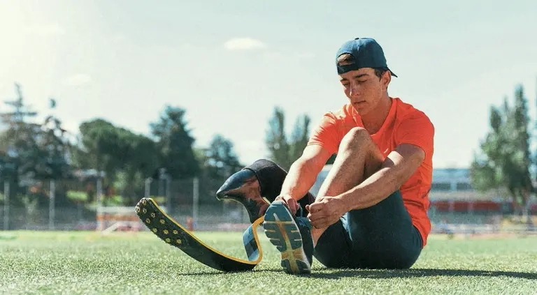 Junger Mann mit in Sportkleidung und mit Cappy sitzt auf dem Boden und bindest sich den Schuh. Am rechten Bein hat er eine Unterschenkelprothese