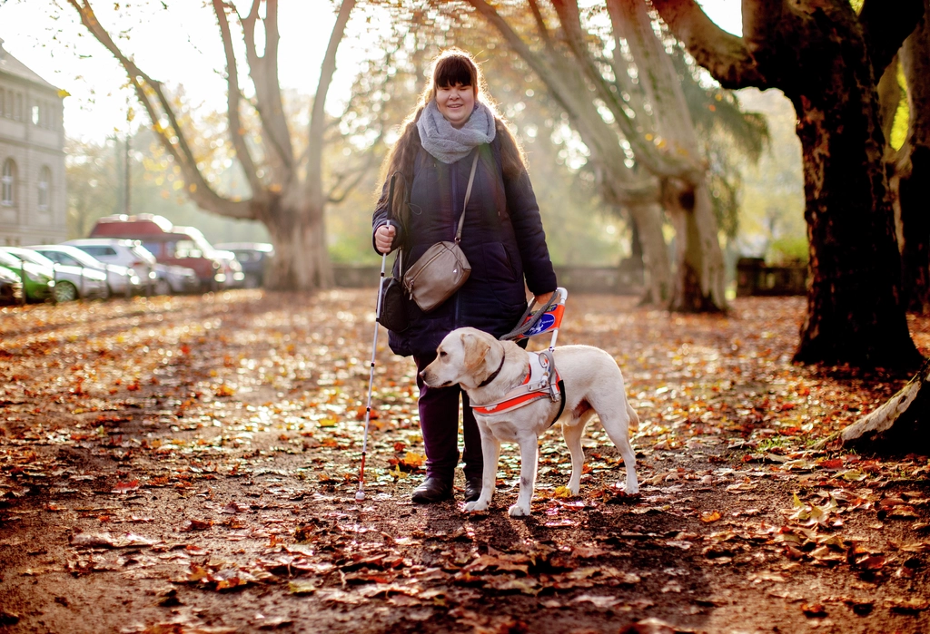 Ein Frau steht mit ihrem Blindenhund auf einem Fußweg.