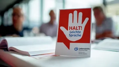 Auf einem Tisch steht ein Schild mit einer warnenden Hand und dem Hinweis: Halt! Leichte Sprache. Im Hintergrund sind unscharf drei Personen zu sehen.