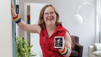 Natalie Dedreux steht in einem roten Jumpsuite in einem Wohnzimmer und lacht in die Kamera. Sie hält sich mit der rechten Hand an der Wand fest.