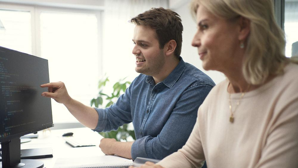 Ein junger Mann und eine blonde Frau sitzen gemeinsam vor einem Computerbildschirm und arbeiten an einem Code.