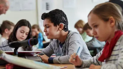 Junge mit einem Hörgerät sitzt in einer Schulklasse. Seine Mitschülerin neben ihm hat ein Mikrophon vor sich stehen über das die Wortbeiträge fü rden Jungen verstärkt werden.