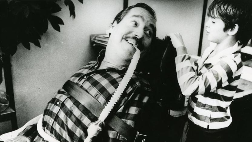 Ein Mann mit Beatmungsschlauch im Rollstuhl schaut lächelnd zu einem Jungen 