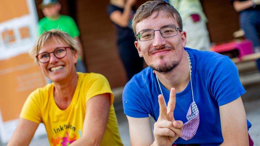 Eine Frau und ein Mann mit geistiger Behinderung sitzen lächelnd nebeneinander. er macht ein Victory-Zeichen