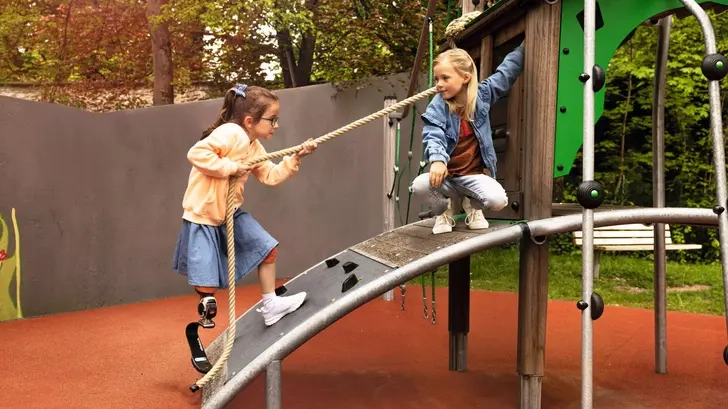 Zwei Mädchen spielen auf einem barrierefreien Spielturm. Ein Mädchen mit Beinprothese erklimmt den Turm gerade, indem sie sich an einem Seil über eine Rampe hochzieht.
