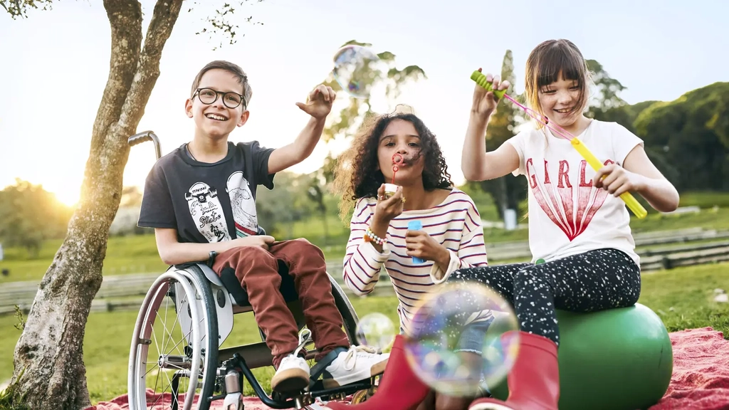 Drei Kinder sitzen in einem Park und pusten Seifenblasen. Ein Kind sitzt im Rollstuhl, ein zweites auf einem Hüpfball. Alle lachen.