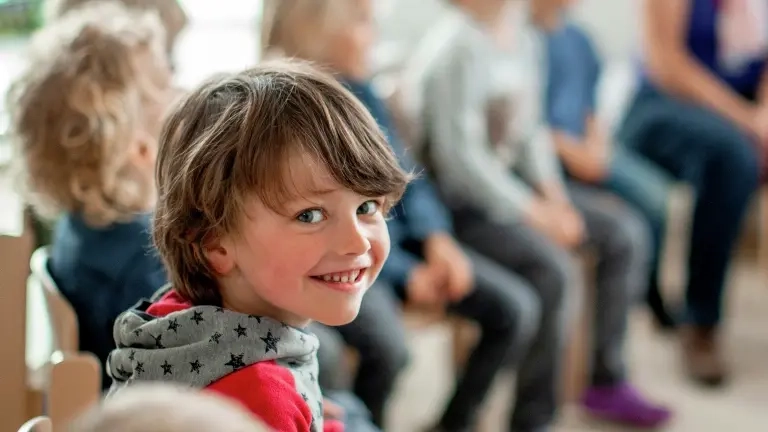 Ein Junge sitzt in einer Kindergartengruppe in einem Stuhlkreis und lächelt über die Schulter in die Kamera.
