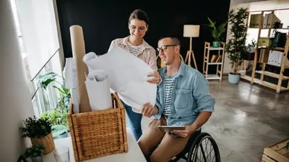 Eine Frau mit einem Plan in der Hand steht neben einem Mann im Rollstuhl. Beide schauen auf den Plan.