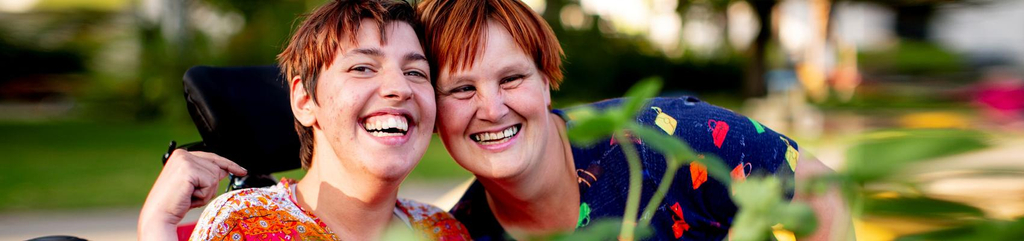 Zwei Frauen in bunt gemusterten Oberteilen sind hinter einem Hochbeet zu sehen. Eine der beiden Frauen sitzt in einem Rollstuhl. Sie stecken die Köpfe zusammen und lachen in die Kamera.