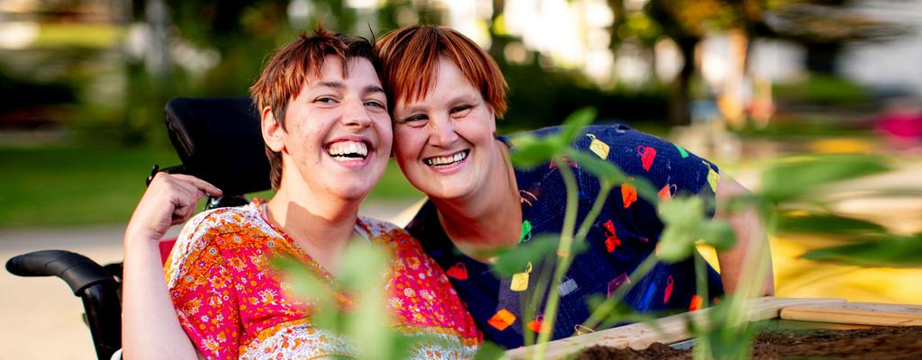 Zwei Frauen in bunt gemusterten Oberteilen sind hinter einem Hochbeet zu sehen. Eine der beiden Frauen sitzt in einem Rollstuhl. Sie stecken die Köpfe zusammen und lachen in die Kamera.