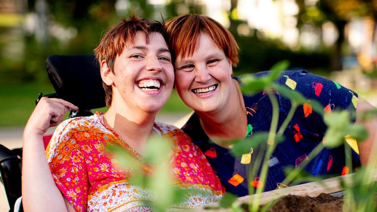 Zwei Frauen in bunt gemusterten Oberteilen sind hinter einem Hochbeet zu sehen. Eine der beiden Frauen sitzt in einem Rollstuhl. Sie stecken die Köpfe zusammen und lachen in die Kamera.