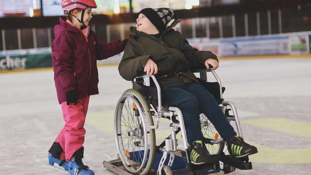 Ein Mädchen auf Schlittschuhen steht auf einer Eisbahn neben einem Jungen im Rollstuhl. Der Rollstuhl steht auf einem Eisgleiter.