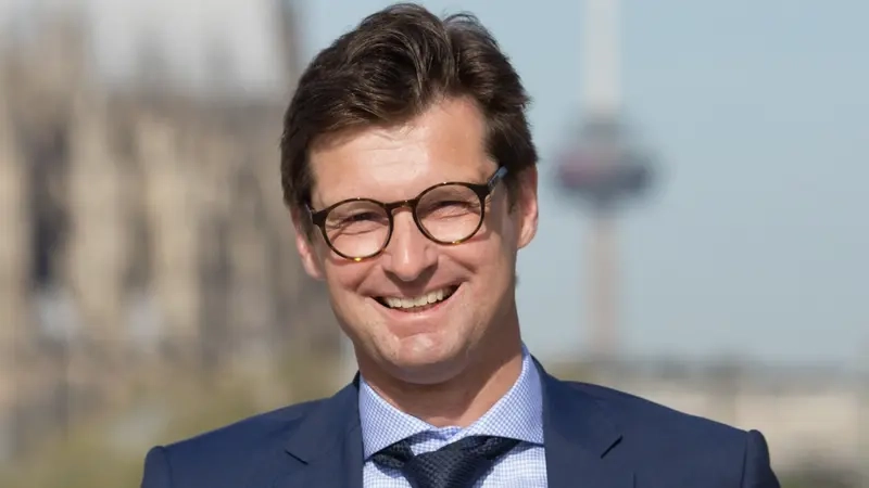 Ein Mann mittleren Alters mit dunkeln Haaren und Brille trägt einen Anzug und schaut in die Kamera.