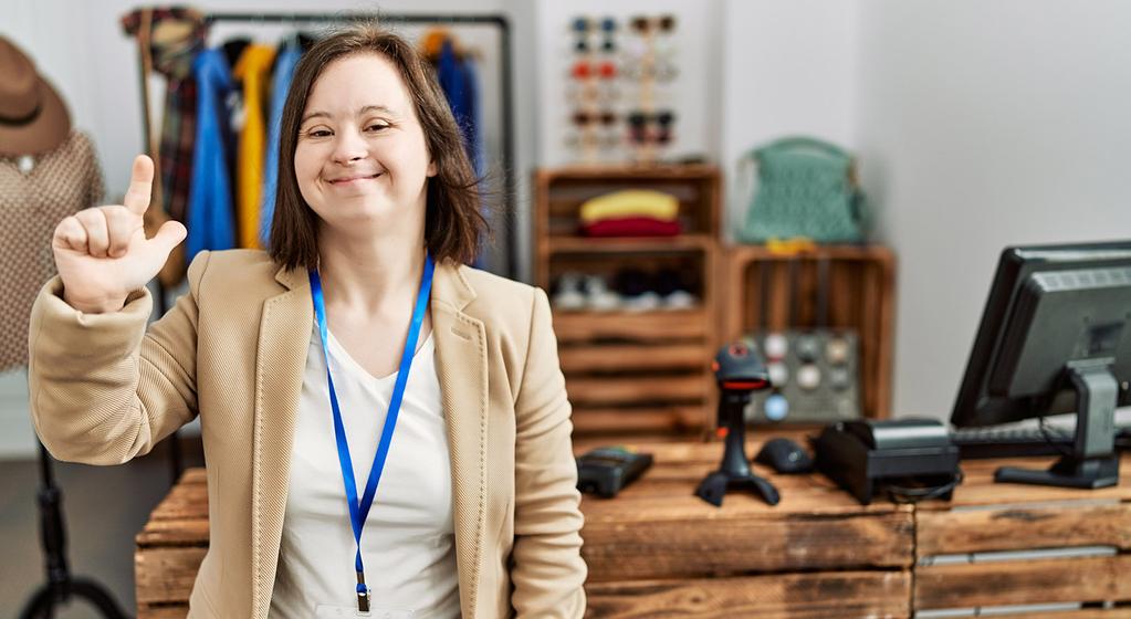Eine junge Verkäuferin mit Down-Syndrom steht in einem Kleidergeschäft. Sie lächelt in die Kamera und zeigt mit der rechten Hand das Symbol für die Zahl zwei.