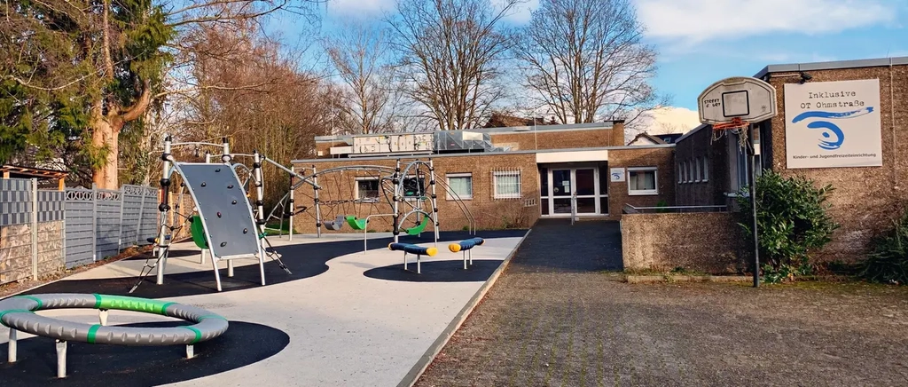 Blick auf ein Gebäude mit Flachdach. Im Vordergrund sind Klettergeräte und ein Trampolin zu sehen. Am Gebäude hängt ein Schild mit der Aufschrift "inklusive OT Ohmstraße"