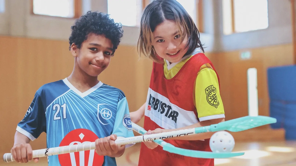 Zwei Jungs unterschiedlicher Hautfarbe balancieren gemeinsam einen Ball zwischen zwei Hockeyschlägern. Sie lachen in die Kamera.