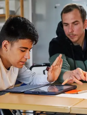 Ein Jugendlicher sitzt an einem Tisch und schaut auf ein Tablet. Neben ihm sitzt Fabio Priano und weist auf etwas auf dem Tablet.