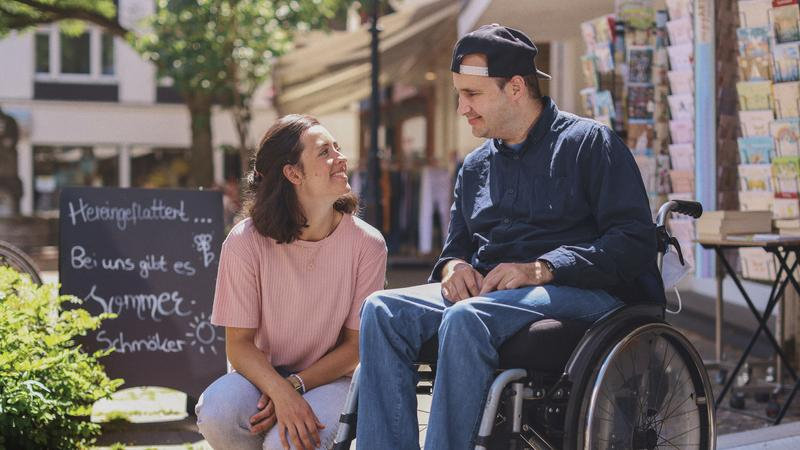 Ein Mann im Rollstuhl sitzt vor einer Buchhandlung. Neben ihm sitzt eine Frau in der Hocke. Die beiden lächeln sich an.