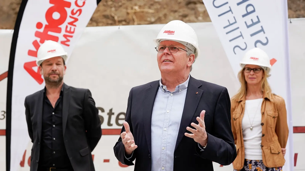 Ein Mann mit grauen Haaren und Brille trägt einen Baustellenhelm und gestikuliert mit den Händen.