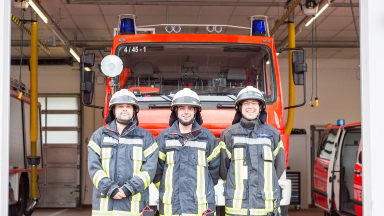 Drei Feuerwehrmänner in Uniform stehen vor einem Feuerwehrauto.