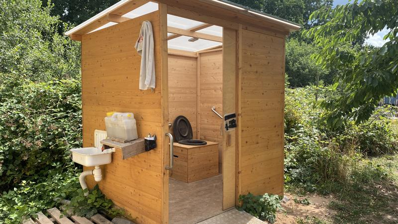 Ein Hütte aus Holz steht in einem Gelände mit Bäumen und Sträuchern. Durch die offene Schiebetür sieht man eine Toilette. Zum Gebäude führt eine Rampe. Außen an der Hütte ist ein Waschbecken angebracht.