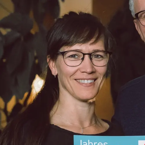 Eine Frau mit langem braunem Haar und Brille und ein Mann mit kurzem grauem Haar stehen Arm in Arm. Gemeinsam halten sie ein Jahreslos in die Höhe.