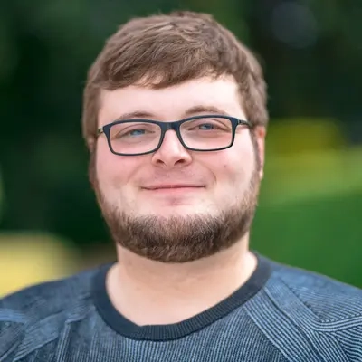 Portrait eines jungen Mannes mit kurzen braunen Haaren und Bart. Er trägt eine Brille und lächelt in die Kamera.