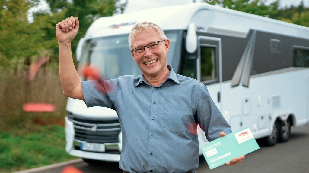 Ein Mann mit kurzem grauen Haar und Brille steht vor jubelnd vor einem Wohnmobil. Er hält ein Glücks-Los in der Hand und es regnet Konfetti.