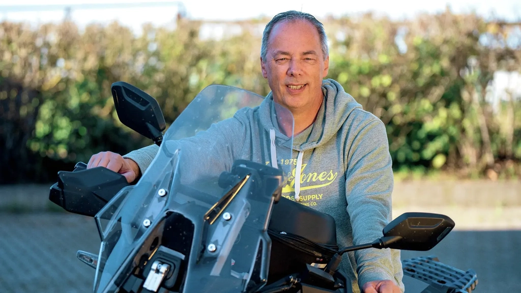 Ein Mann mit kurzem grauen Haar und im Kapuzenpulli sitzt auf einem schwarzen Motorrad.