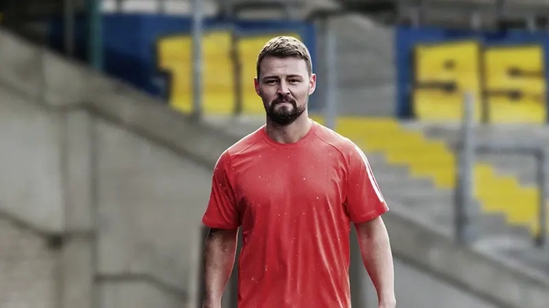 kräftiger junger Sportler mit dunkeln Haaren, kurzem Vollbart, rotem T-Shirt, kurzer Laufhose und Prothese steht auf der Laufbahn und schaut in die Kamera