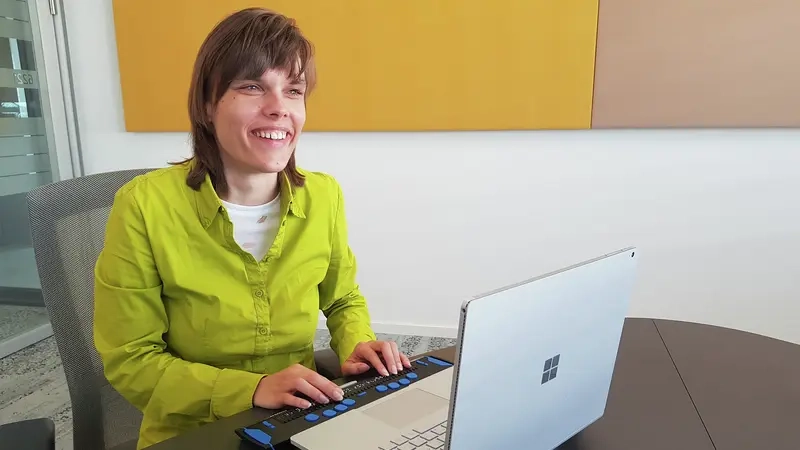 Eine blinde junge Frau sitzt lächelnd vor einem Laptop mit Braillezeile