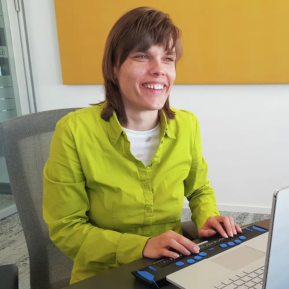 Eine blinde junge Frau sitzt lächelnd vor einem Laptop mit Braillezeile