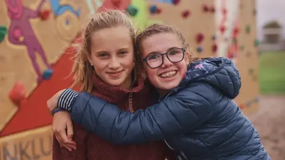 Zwei Jugendliche stehen vor einer Kletterwand. Die eine umarmt die andere.