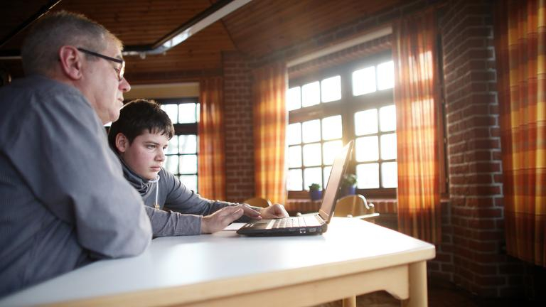 Ein Junge und ein Herr sitzen gemeinsam an einem Tisch und schauen auf einen Laptop.