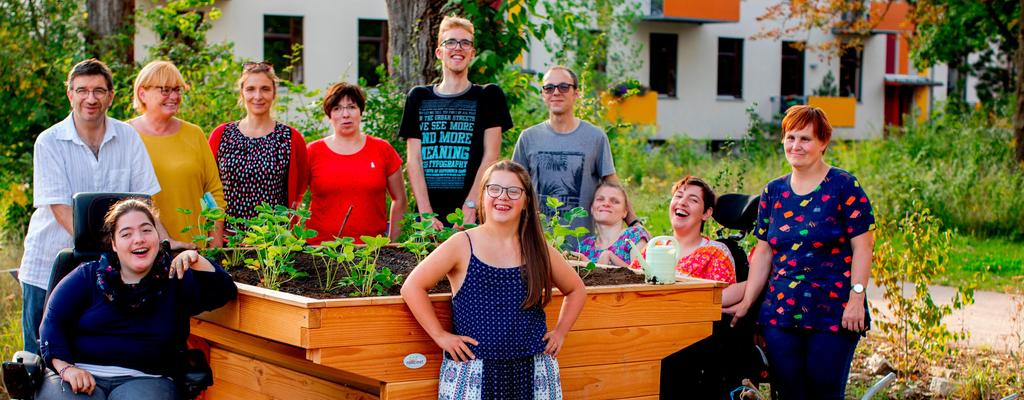Eine Gruppe von Menschen mit und ohne Behinderung steht um ein Hochbeet, das sie gerade bepflanzt haben. Alle lächeln.