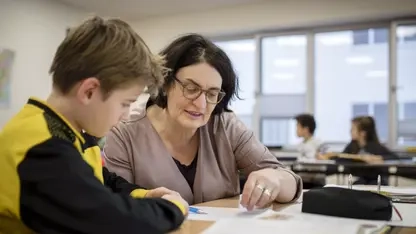 Eine Lehrerin sitzt mit einem Schüler an einem Tisch im Klassenzimmer und erklärt ihm eine Aufgabe.