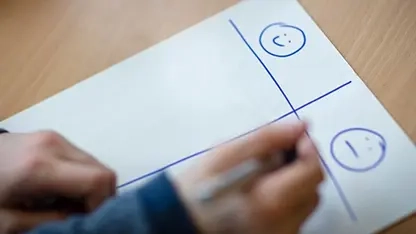 Kinderhände mit einem Stift in der Hand über einem Blatt. Auf diesem Blatt sieht man eine Tabelle mit einem lachenden und einem weinenden Smiley.