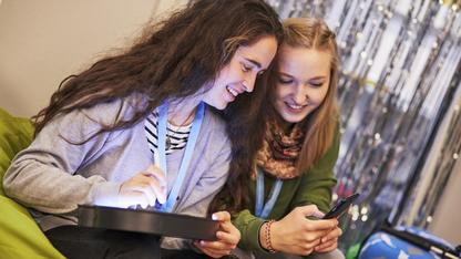 Zwei jugendliche Mädchen mit Handy und Tablet schauen lächelnd aufs Handy