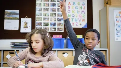 Ein Mädchen und ein Junge sitzen nebeneinander an einem Tisch in einem Klassenzimmer. Das Mädchen schreibt in ihr Heft, der Junge zeigt mit gehobenen Arm auf. 