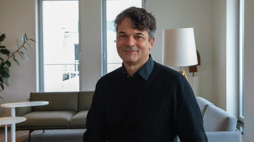 Robert trägt einen schwarzen Pullover und ein schwarzes Hemd. Er steht vor einem Sofa im Aufenthaltsraum und lächelt in die Kamera.