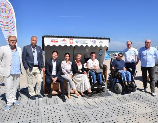 Frauen und Männer stehen neben oder sitzen im inklusiven Strandkorb. Zwei Menschen sind Rollstuhlfahrer.