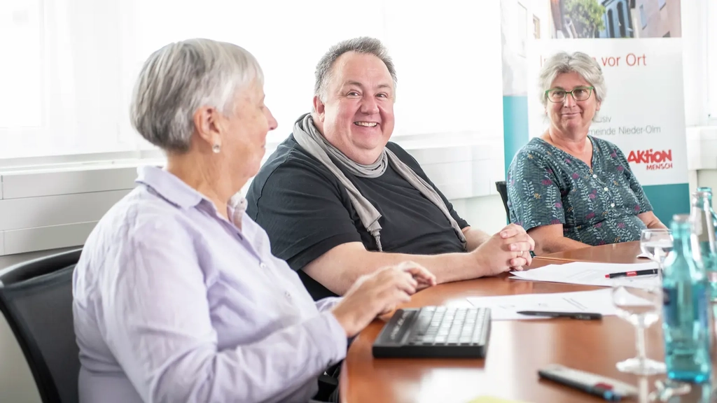 Jörg Greis sitzt am Konferenztisch, links und rechts von ihm sitzt jeweils eine Frau
