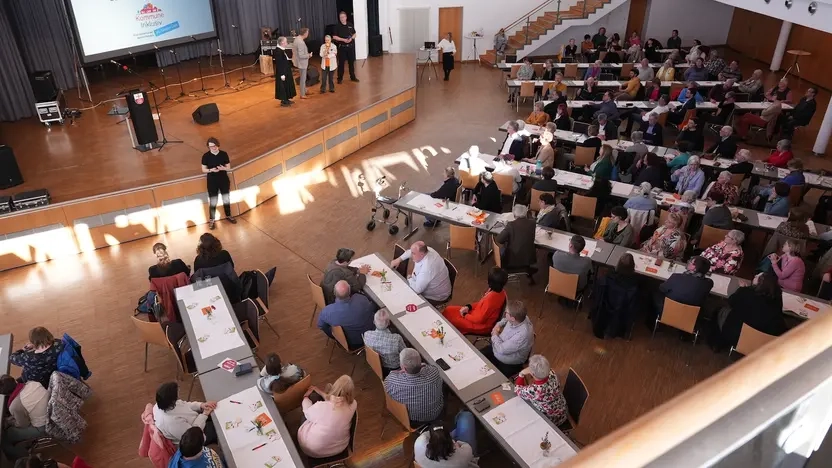 Ein großer Saal, wo viele Menschen an Tischen sitzen und zur Bühne schauen