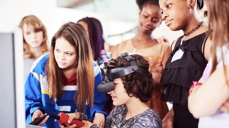 Eine junge Frau mit Virtual-Reality-Brille sitzt vor einem Computerbildschirm und hat einen Joystick in der Hand.  Andere junge Frauen schauen zu.
