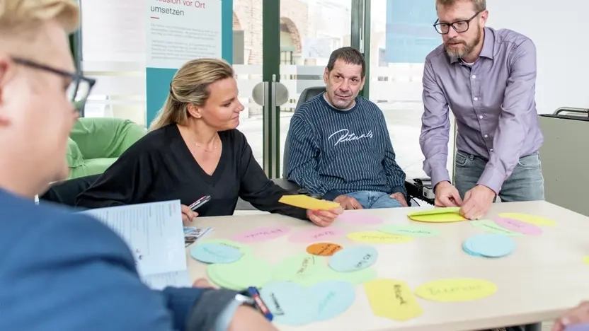 Menschen mit und ohne Behinderung sitzen an einem Tisch und schreiben sich Stichwörter auf ovale, bunte Kärtchen auf