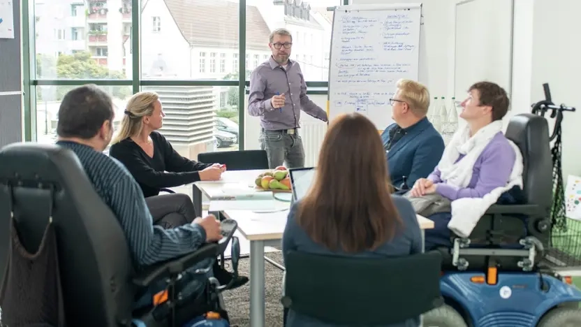 Menschen mit und ohne Behinderung arbeiten zusammen in einem Workshop, eine Person steht an einer Flipp-Chart