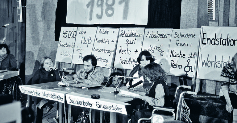 drei Menschen mit Behinderung auf einem Podium mit Mikros. Hinter ihnen stehen Transparente und Schilder mit Sprüchen zur gesellschaftlichen Situation von Menschen mit Behinderung