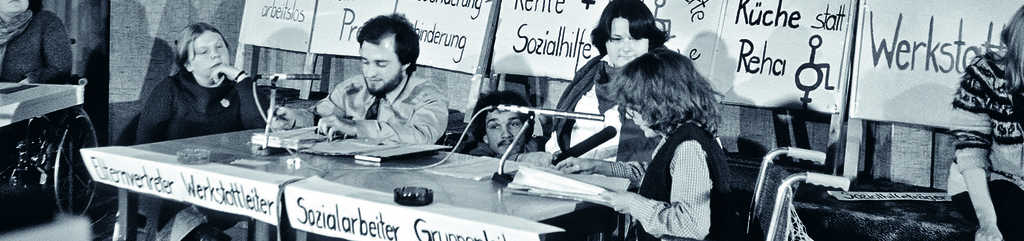 drei Menschen mit Behinderung auf einem Podium mit Mikros. Hinter ihnen stehen Transparente und Schilder mit Sprüchen zur gesellschaftlichen Situation von Menschen mit Behinderung