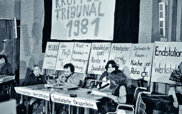 drei Menschen mit Behinderung auf einem Podium mit Mikros. Hinter ihnen stehen Transparente und Schilder mit Sprüchen zur gesellschaftlichen Situation von Menschen mit Behinderung